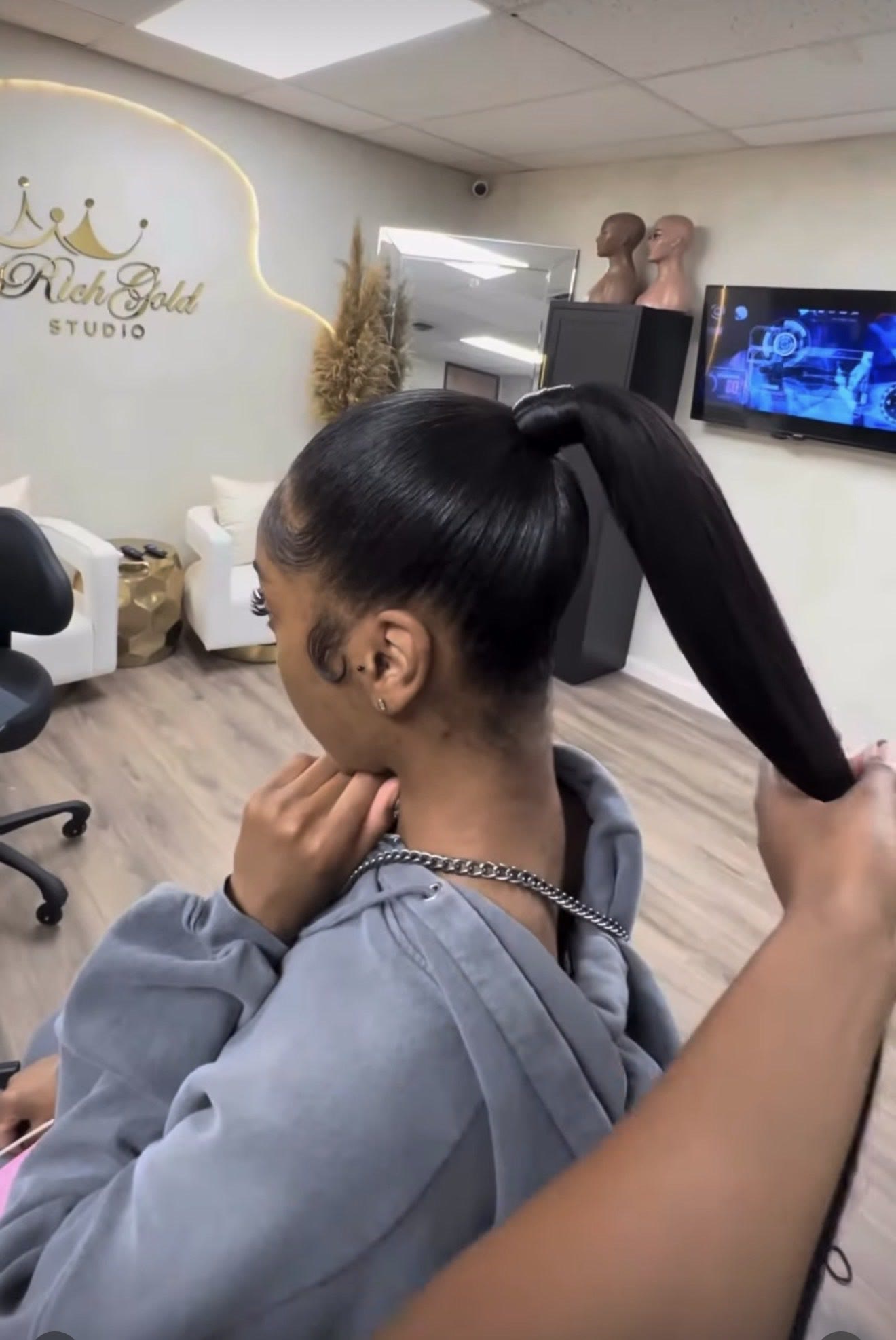 A stylist finishing a long sleek ponytail on a client, with the lit RichGold Studio sign on the wall behind them.