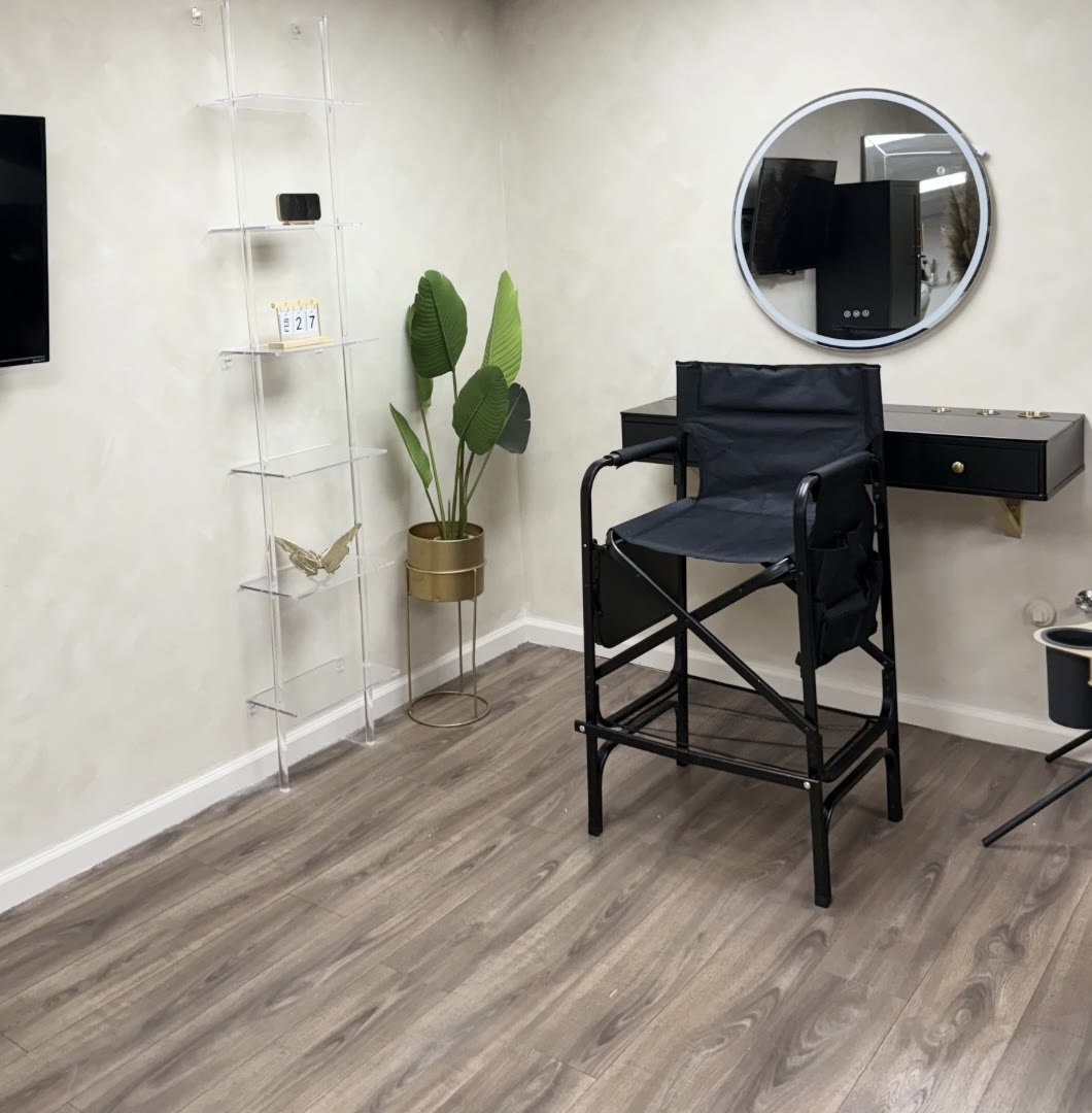 The makeup station: a tall directors chair at a black vanity under a round lit mirror, beside a clear shelving unit and a plant.
